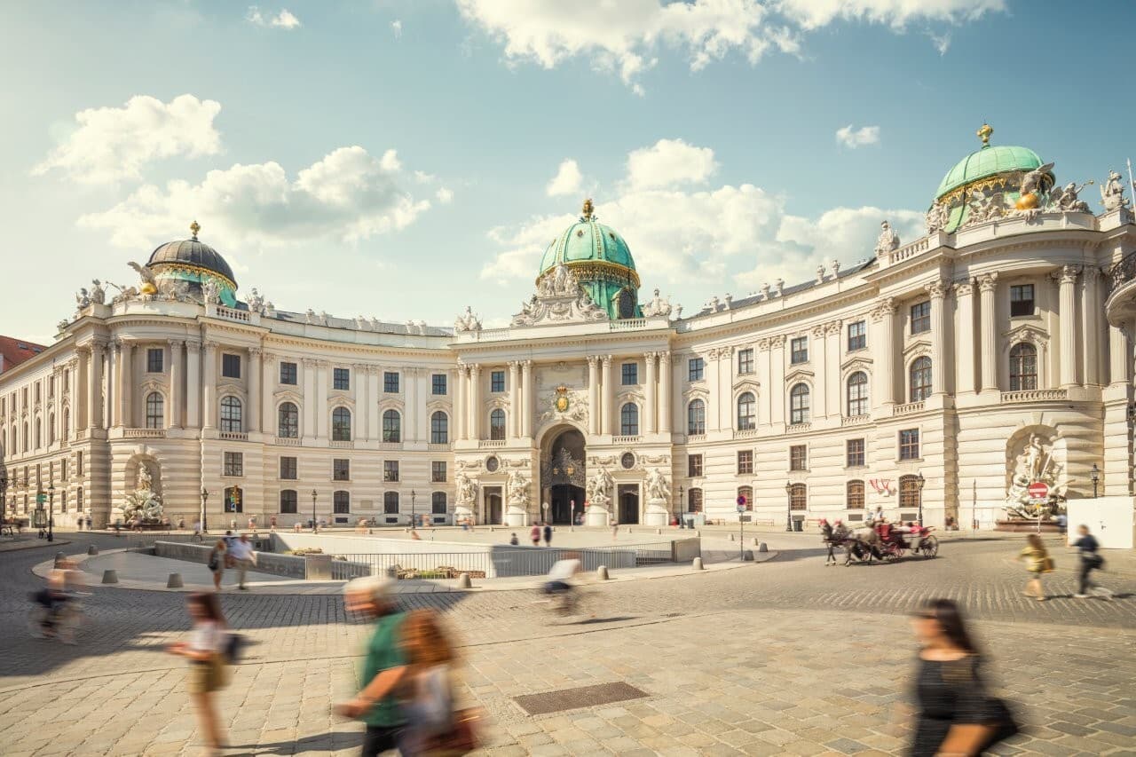 The Hofburg's curved white facade and green domes on Michaelerplatz, passers-by blurred in the sun