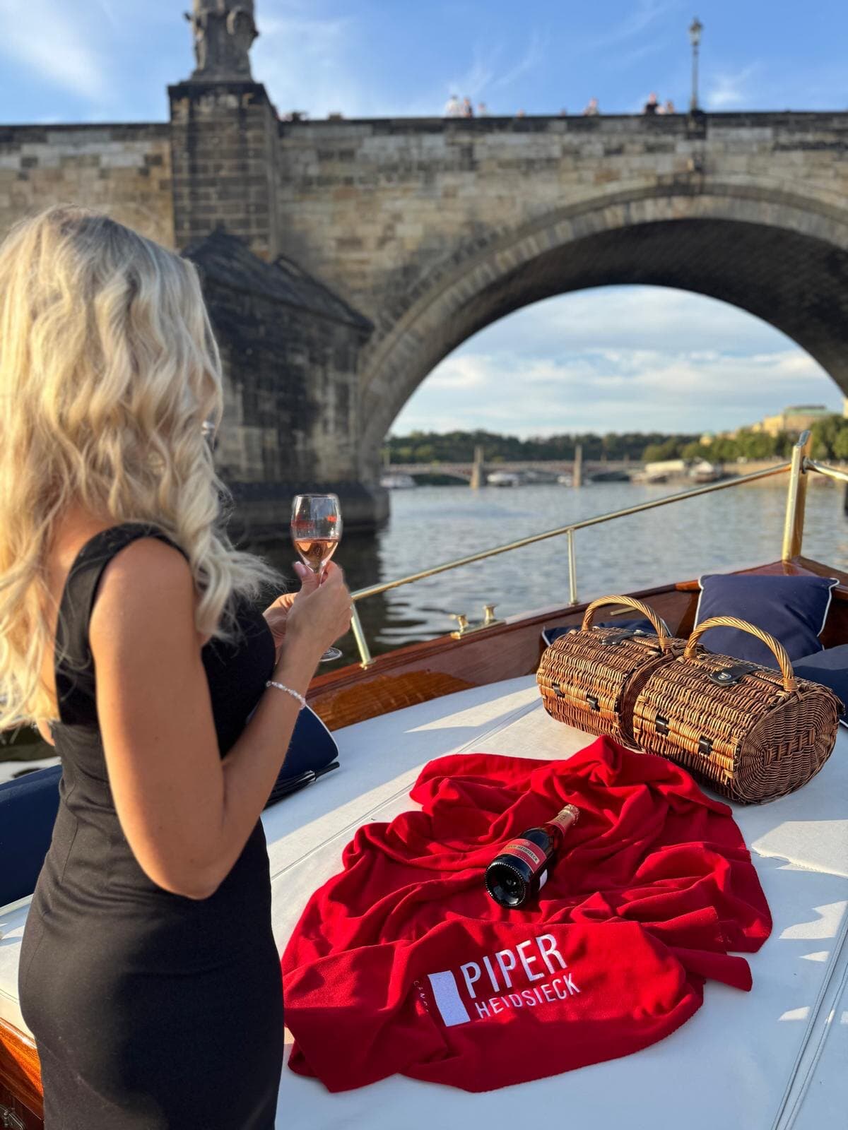 Woman in a black dress holding a glass of rosé on the deck of a wooden boat passing under a stone bridge, a bottle of Piper-Heidsieck on a red blanket beside wicker baskets