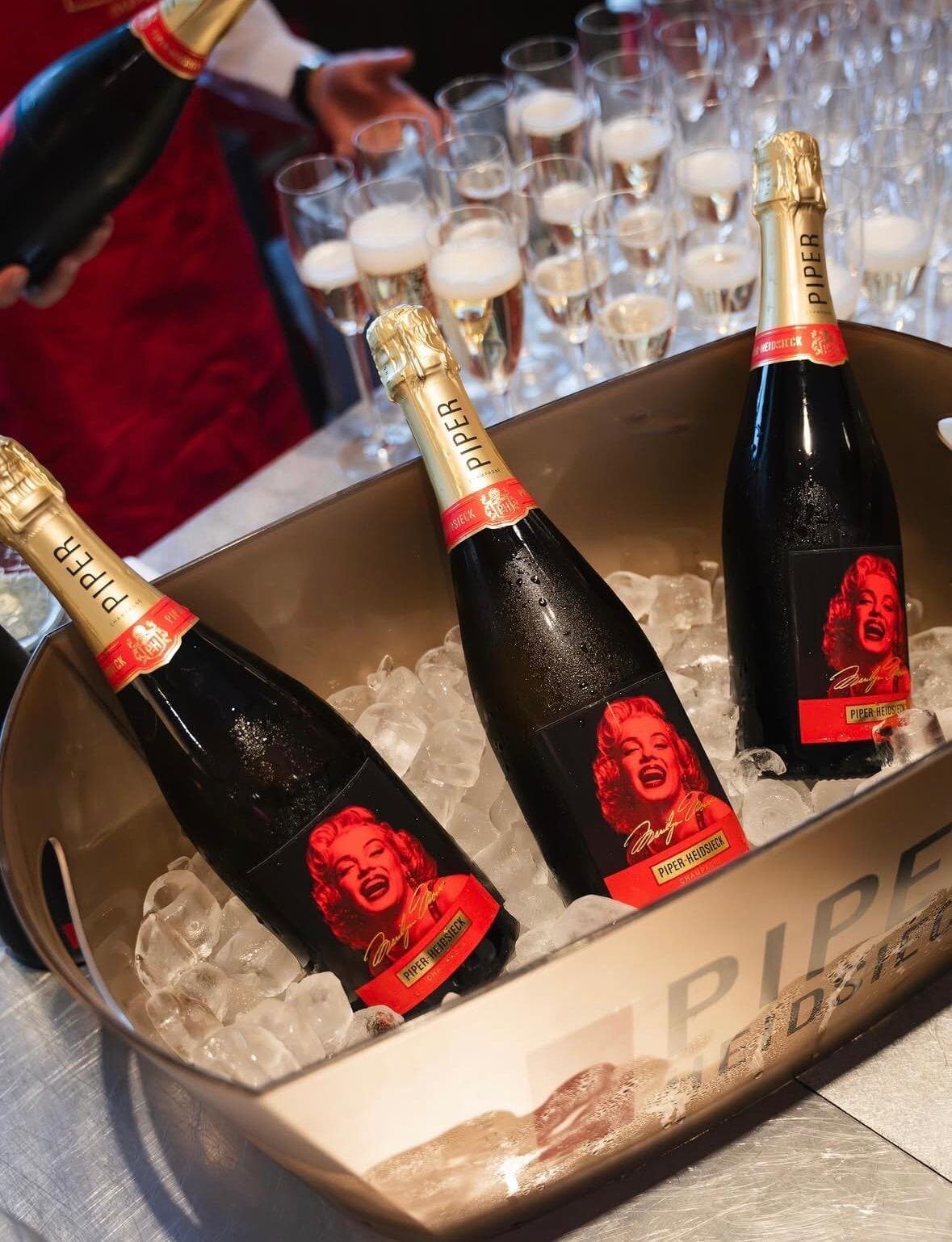 Three bottles of Piper-Heidsieck with Marilyn Monroe labels on ice, rows of poured flutes behind