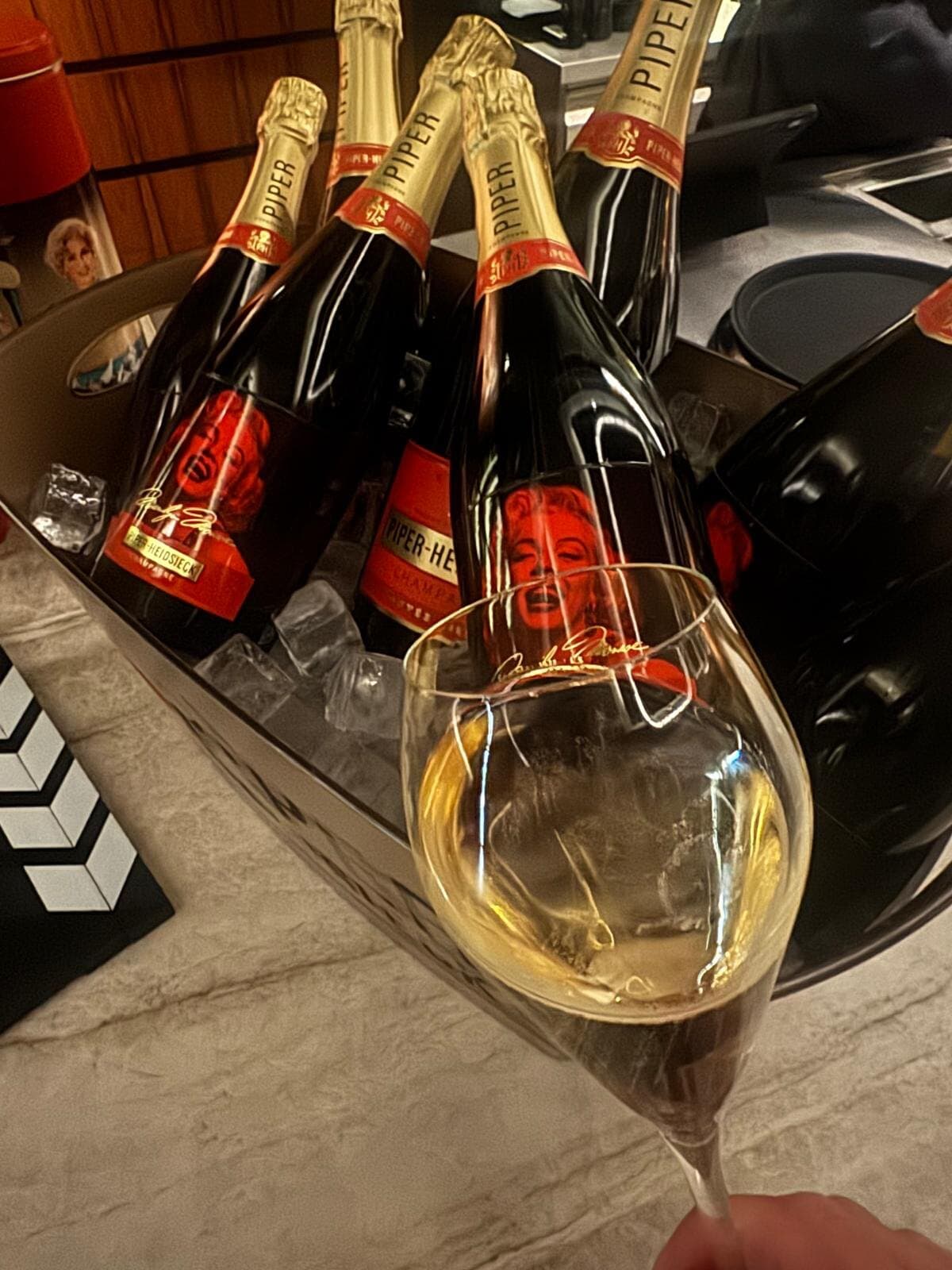 Glass of champagne raised in front of Piper-Heidsieck bottles with Marilyn Monroe labels in an ice bucket