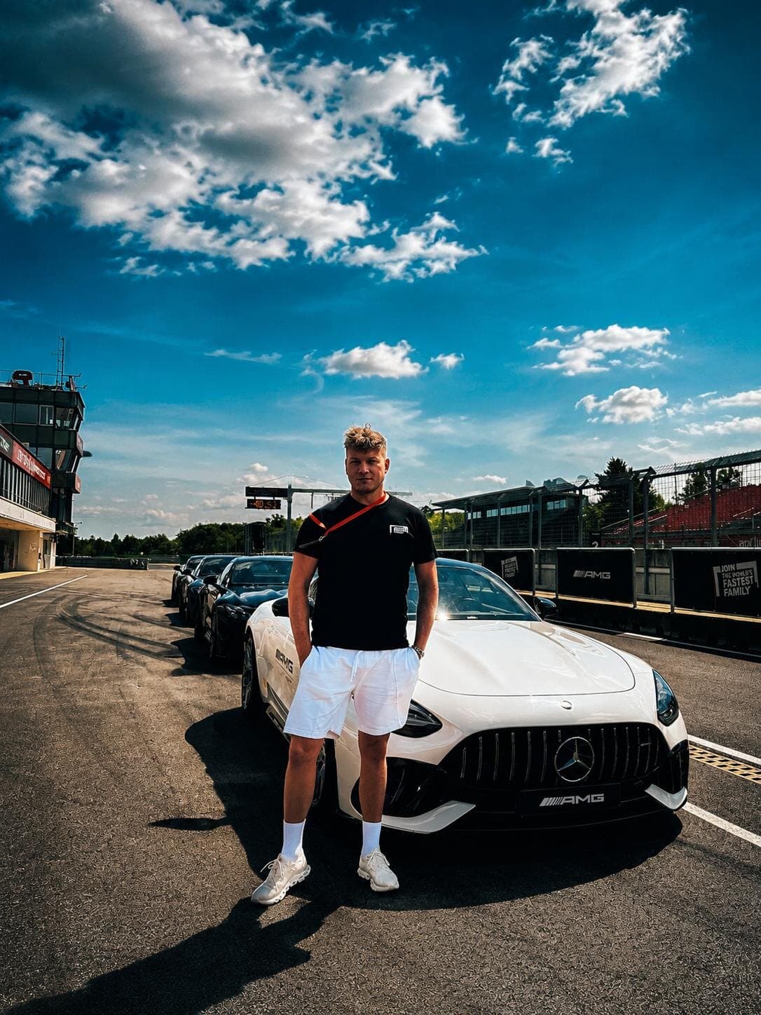 Man standing in front of a white Mercedes-AMG in a race track pit lane, more cars lined up behind him