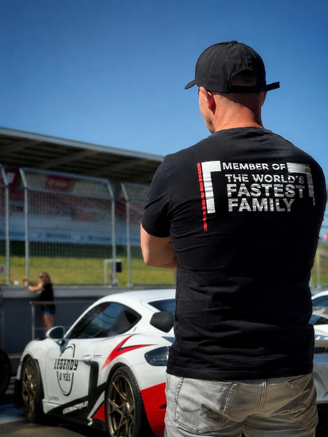 Man seen from behind in a black T-shirt reading "Member of the world's fastest family", white and red sports cars in front of the grandstand