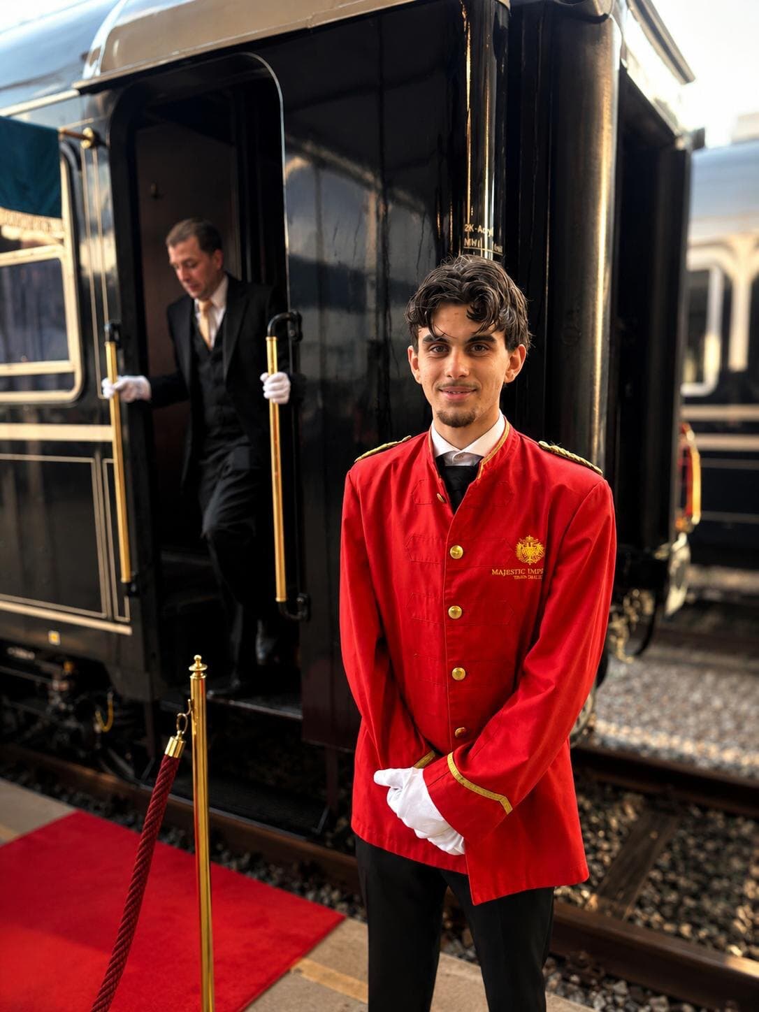 Steward in a red uniform jacket and white gloves on a red carpet beside a vintage black train carriage, a second attendant stepping down from the door