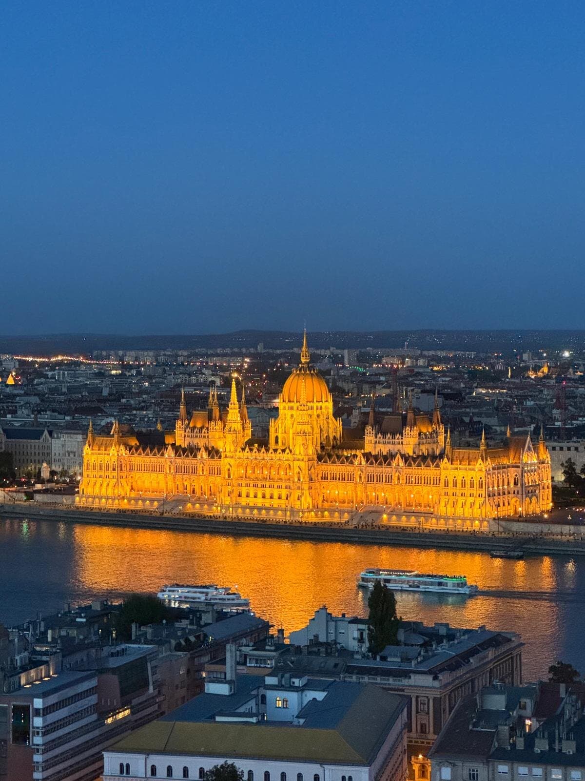 The floodlit Hungarian Parliament reflected in the Danube at dusk, two river boats passing