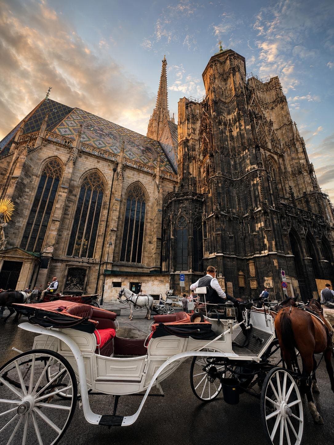 A white horse-drawn carriage with red seats in front of St. Stephen's Cathedral and its patterned tile roof at sunset
