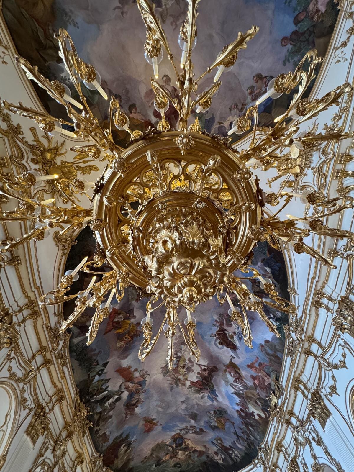Looking straight up at a gilded chandelier against a white and gold stucco ceiling with frescoes
