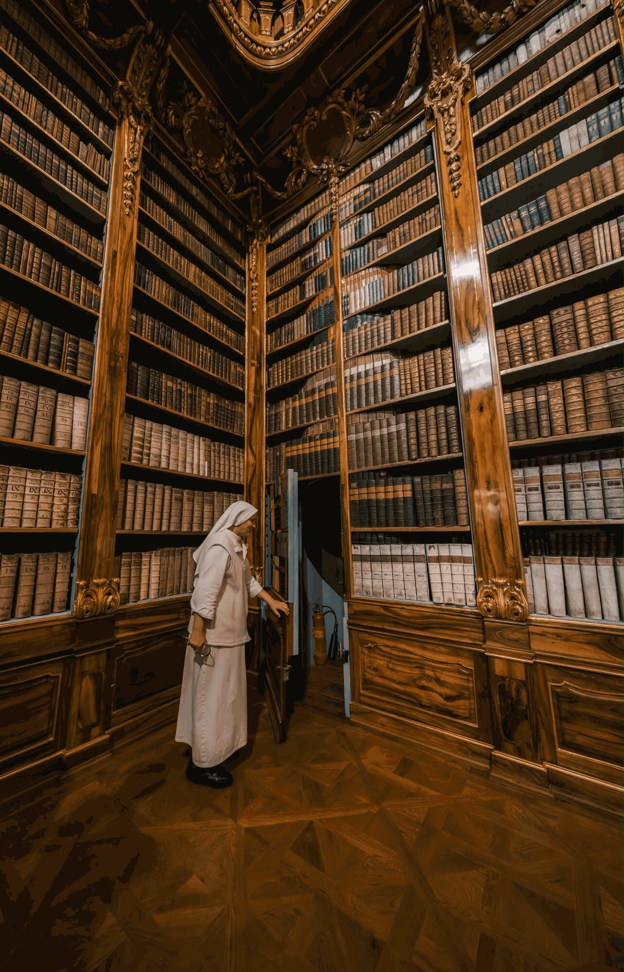 A nun opens a hidden door between the bookcases of a baroque library