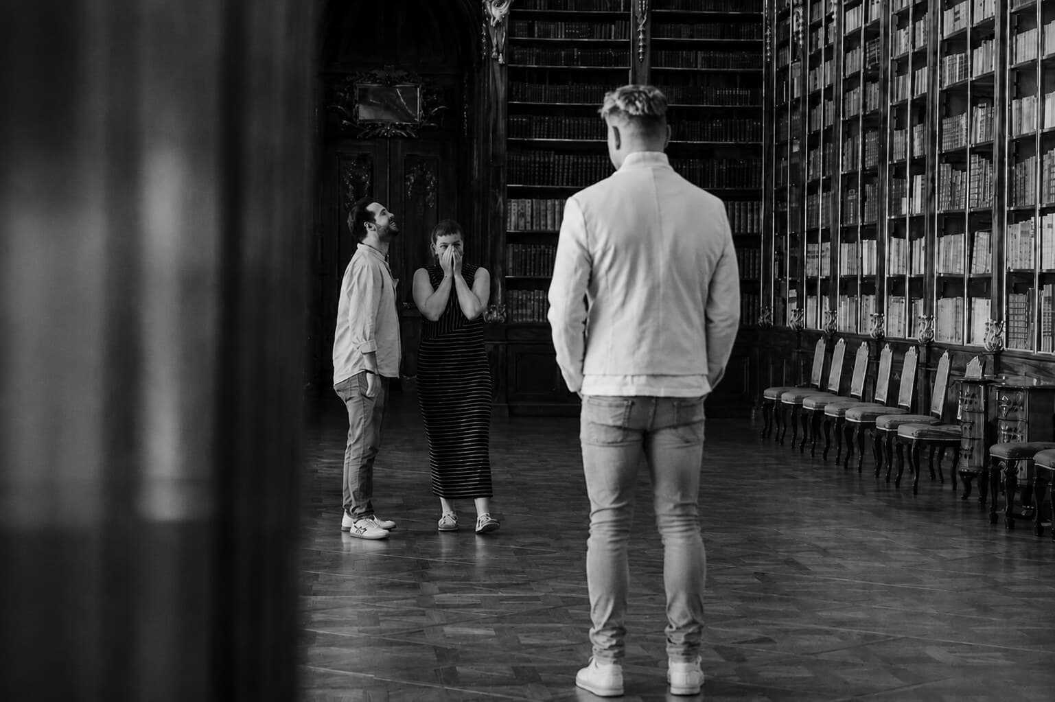 Black-and-white photo of a library hall lined with tall bookcases: a man in a white jacket faces a couple, the woman covering her face with her hands