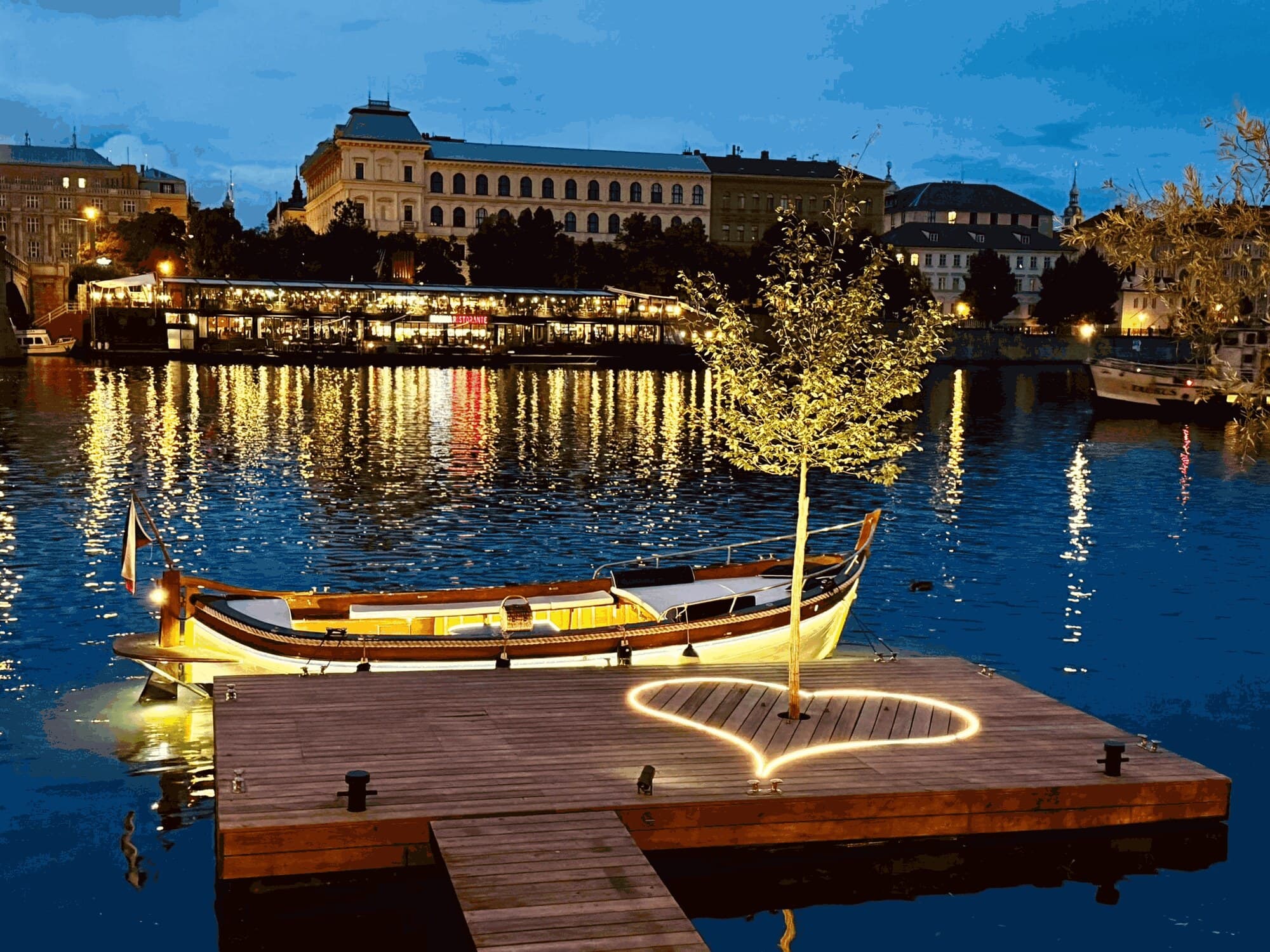 A wooden pier on the river at dusk with a glowing heart of lights around a young tree, a varnished boat moored alongside and lit buildings across the water