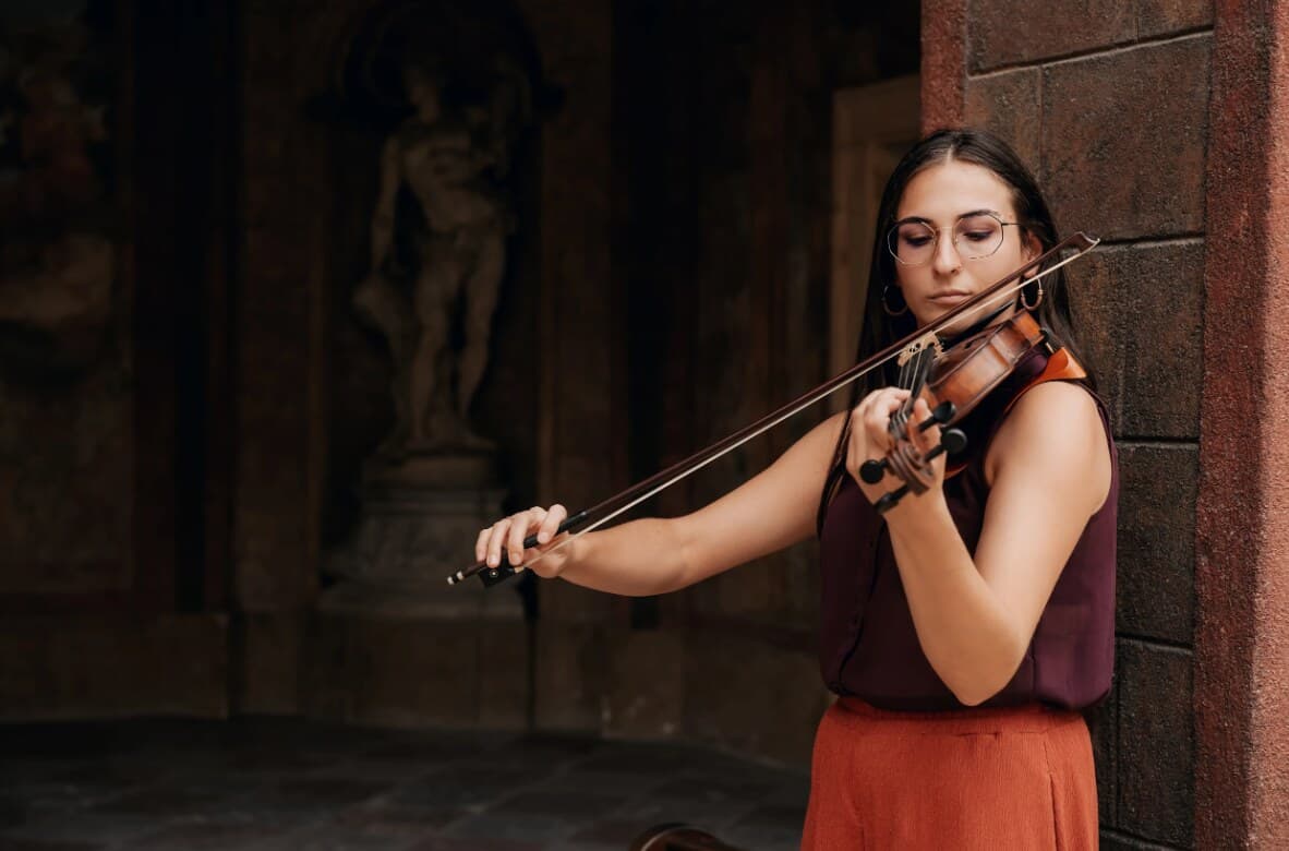 Violinist playing beside a stone wall, a statue in a dim niche behind her